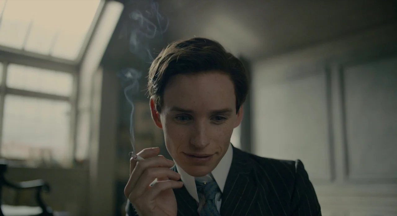 The Danish Girl (2015) movie still: closeup — A man in a 1920s-style suit sits indoors near a large window. He holds a lit cigarette between his…