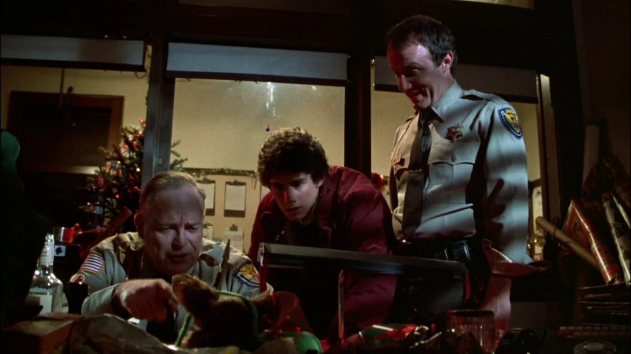 Gremlins (1984) movie still: medium — A dimly lit local police station office interior during the holiday season. Two uniformed officers and a…