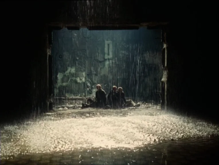 Stalker (1979) movie still: wide — Three people sit on the ground inside a dark, concrete structure with crumbling walls. Rain pours down in a…