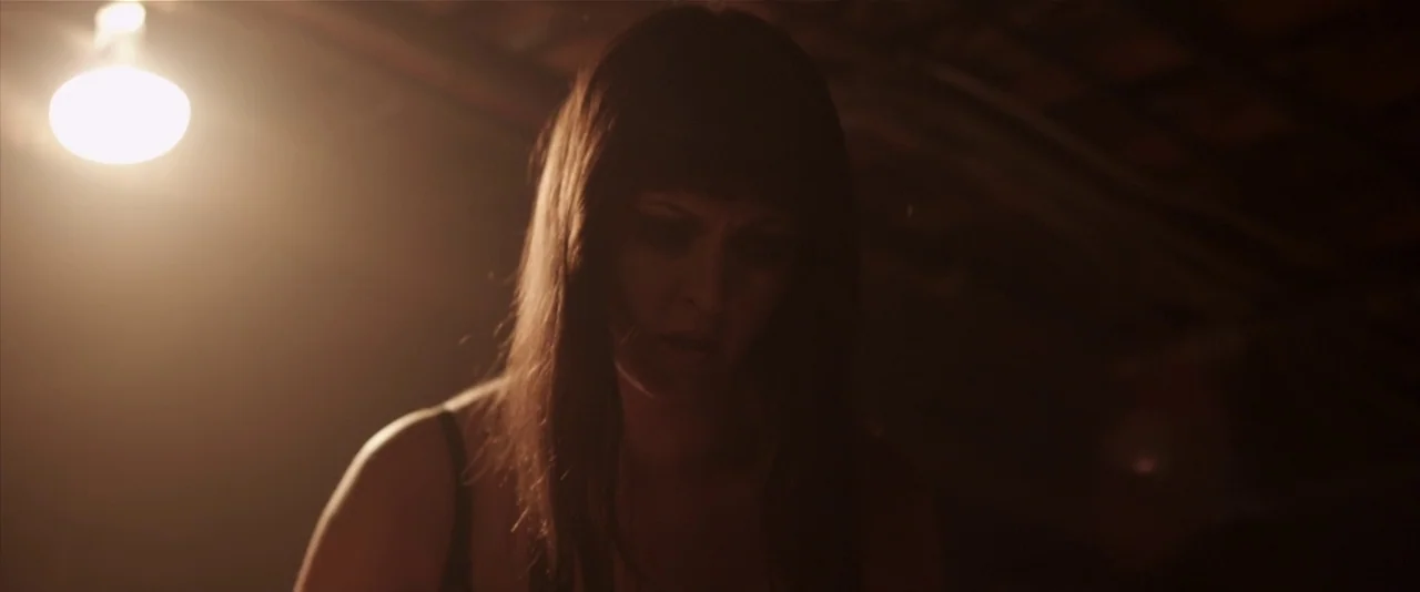 American Mary (2012) movie still: medium — A dark, cramped space with rough overhead wooden rafters visible in the shadows. A young woman with dark…
