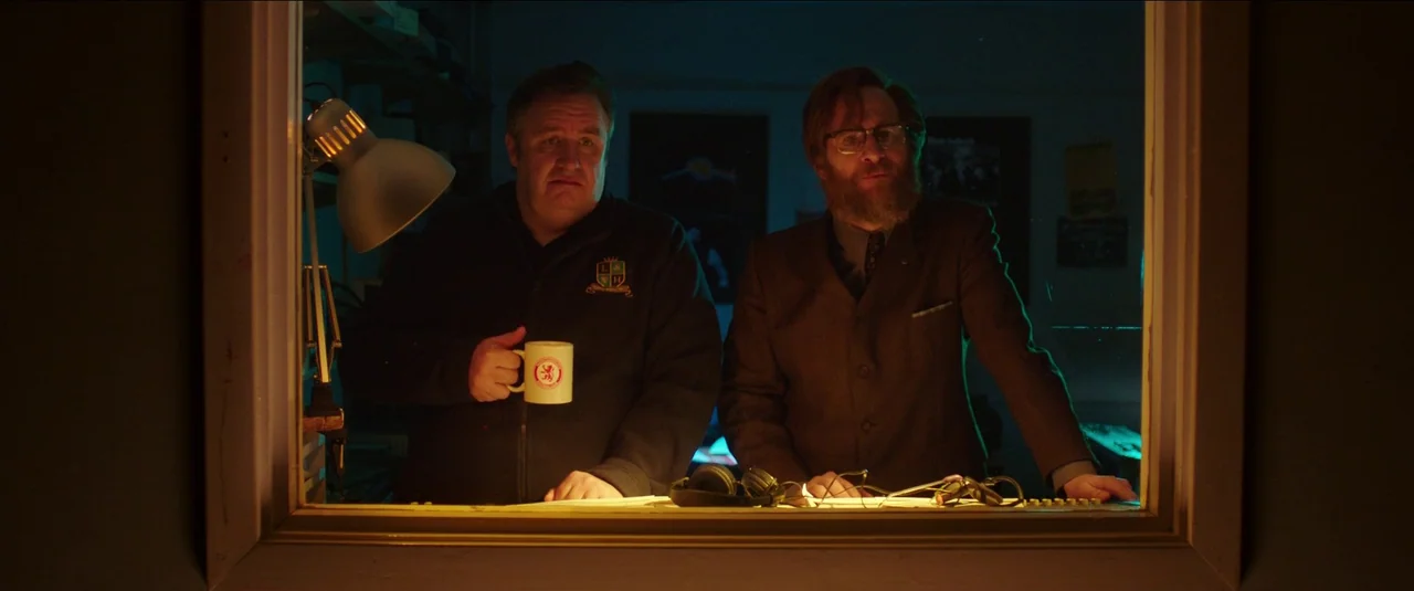 Anna and the Apocalypse (2018) movie still: medium — Two men stand inside a dark broadcast booth partitioned by a wooden frame window. They face…