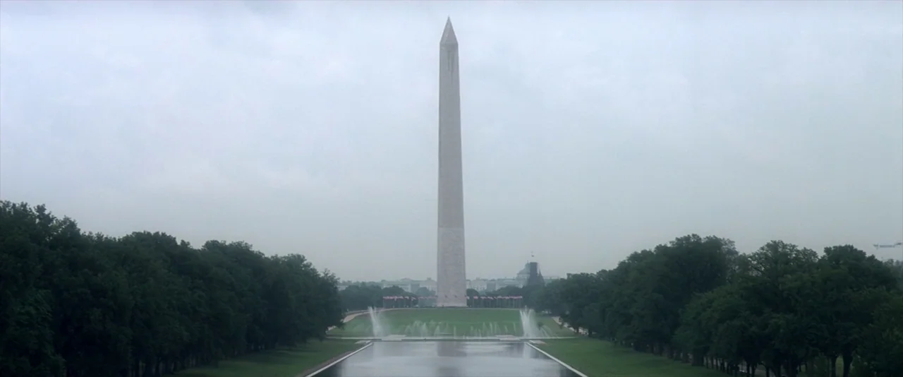 JFK (1991) movie still: establishing — The Washington Monument stands prominently at the center of the frame under grey skies. Rows of trees flank…