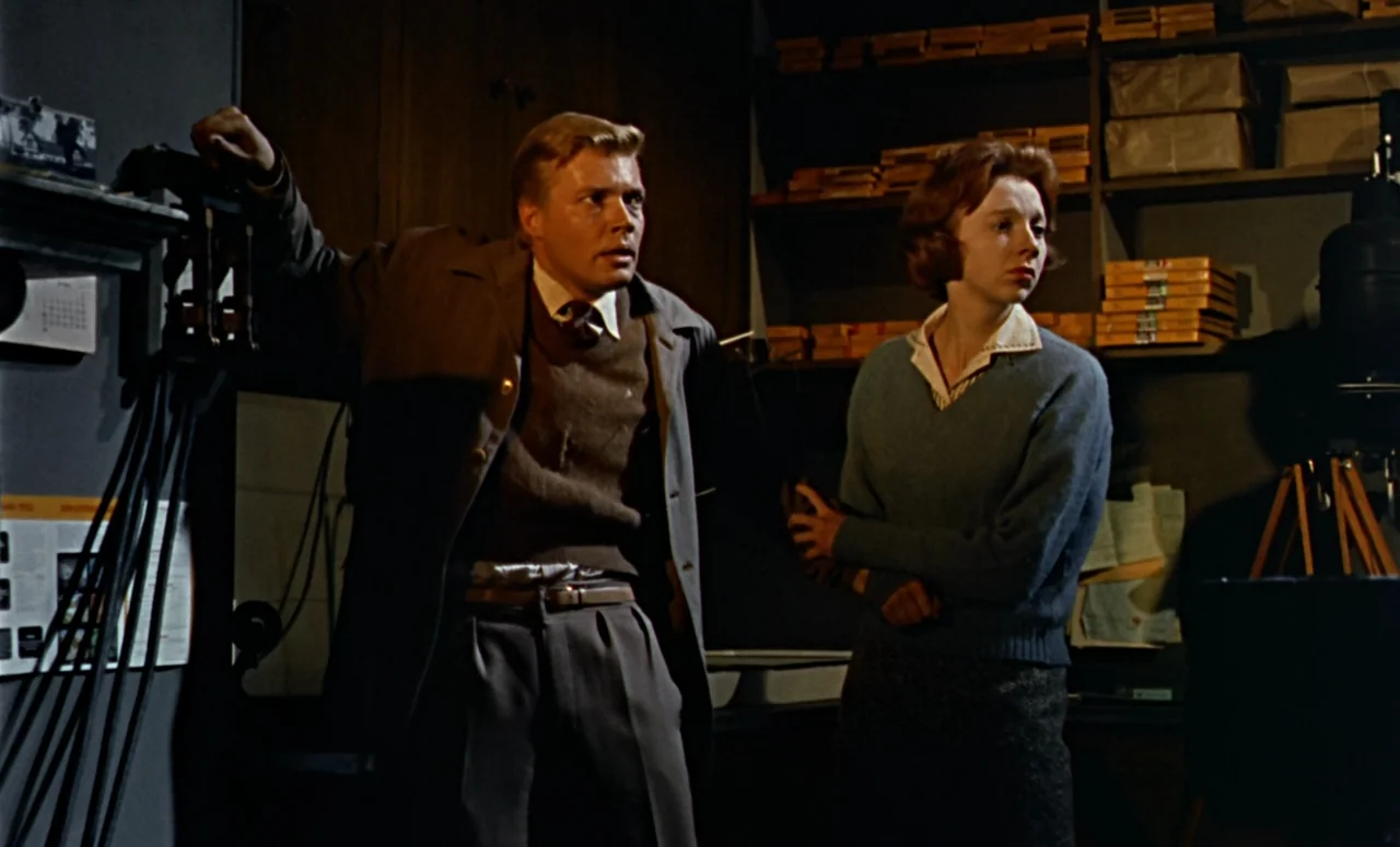 Peeping Tom (1960) movie still: medium — A dark, cluttered archive room contains shelves packed with stacked, wrapped paper bundles. A young man and…