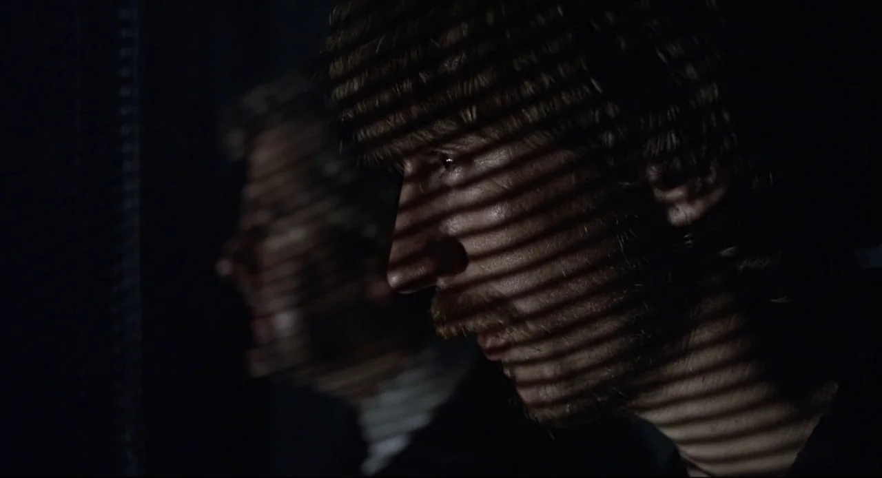 Naked (1993) movie still: closeup — Two men stand in a dark room illuminated by light filtering through blinds. The men gaze out of a window with…