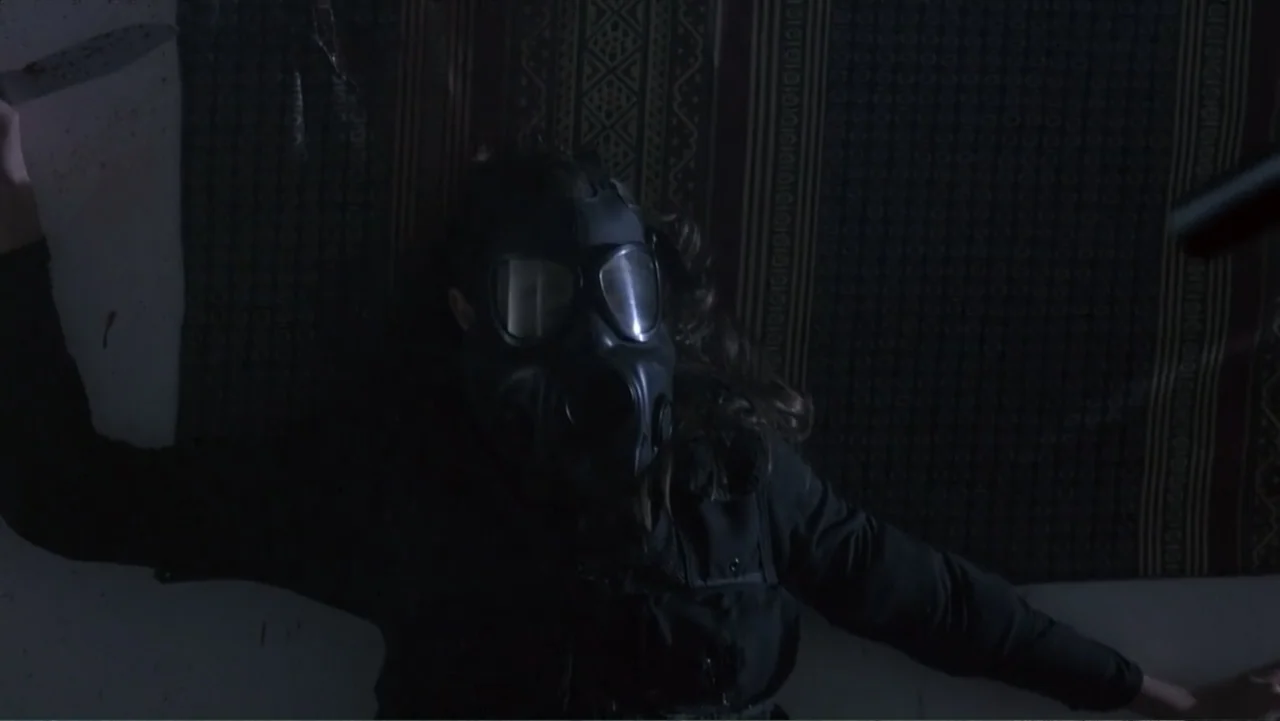 Crisis Point (2012) movie still: medium — A small dark interior features a wall draped with a patterned fabric backdrop. A figure wearing a gas mask…