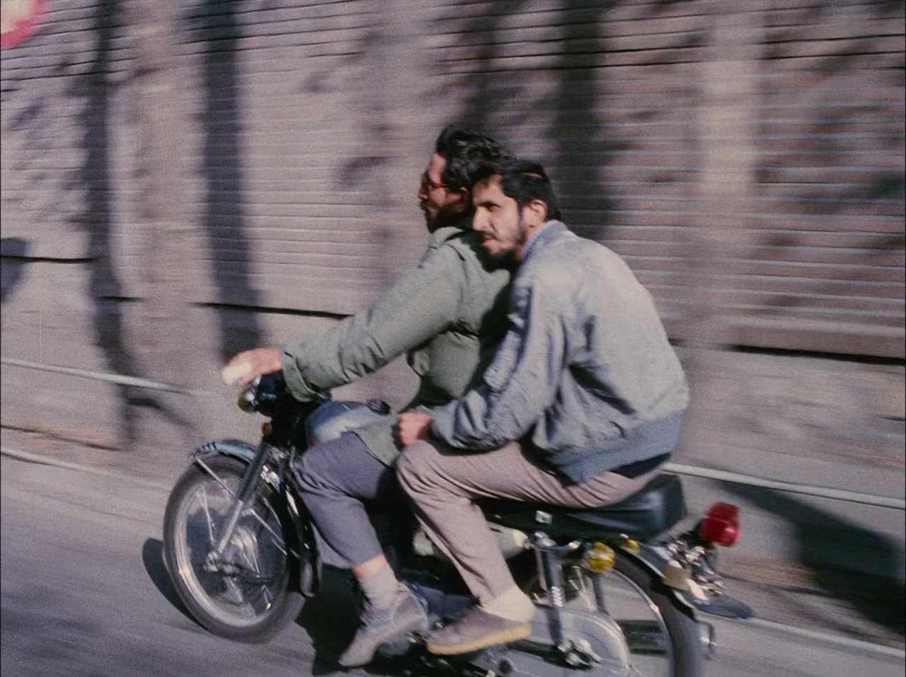 Close-Up (1990) movie still: medium — Two men ride a motorcycle along a road beside a long brick wall. The motion blur indicates speed while the…