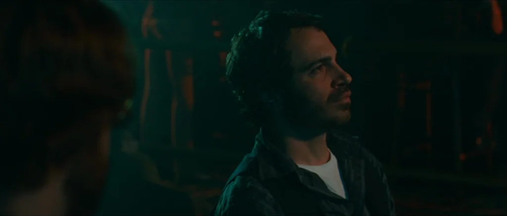 Away We Go (2009) movie still: medium — A dark indoor space with teal ambient lighting and indistinct shapes in the background. A man sits facing…