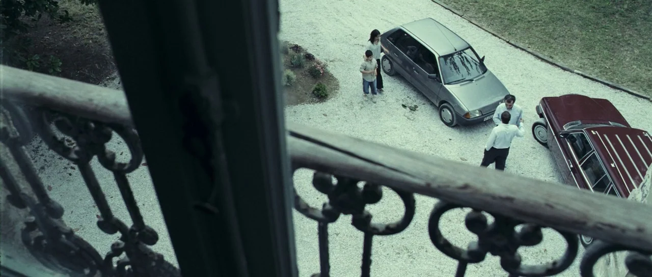 The Orphanage (2007) movie still: wide — A gravel driveway is viewed from a high-angle perspective behind a wrought iron railing. Two men face each…
