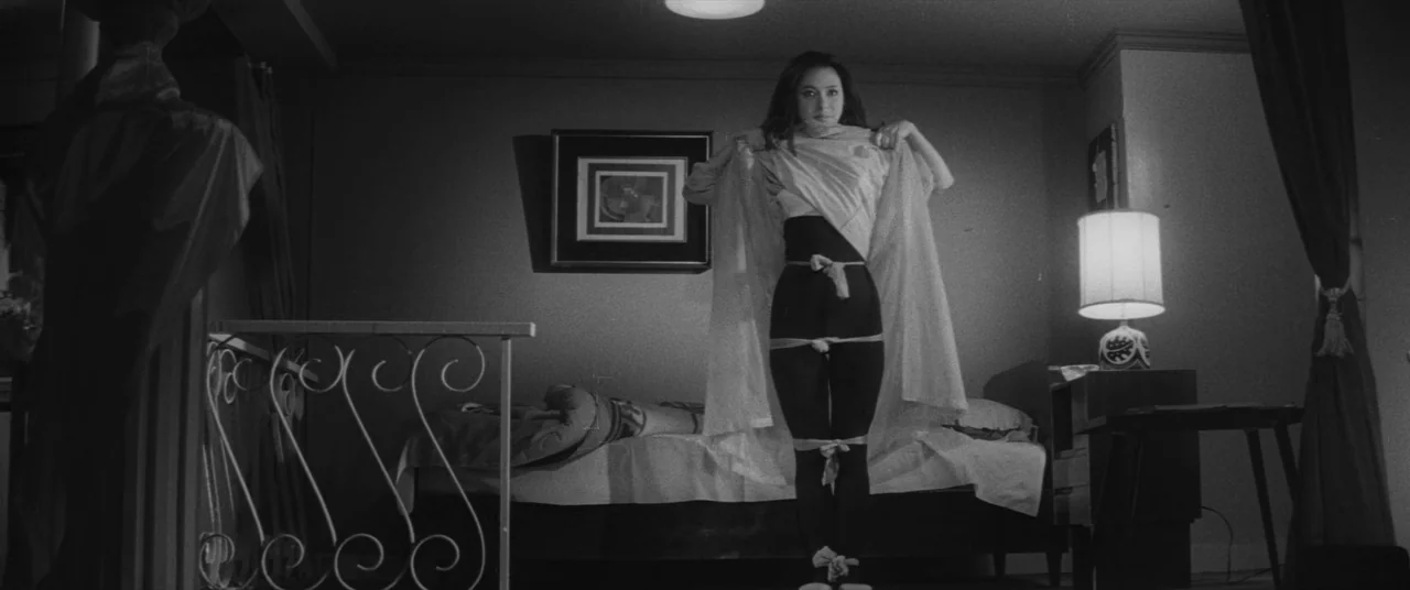 Carmen From Kawachi (1966) movie still: fullbody — A dark, sparsely furnished room contains a bed and a decorative lamp on a stand. A young woman in…