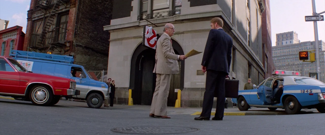 Ghostbusters (1984) movie still: wide — An exterior city scene features a brick firehouse building with a ghost logo sign. Two men in business suits…