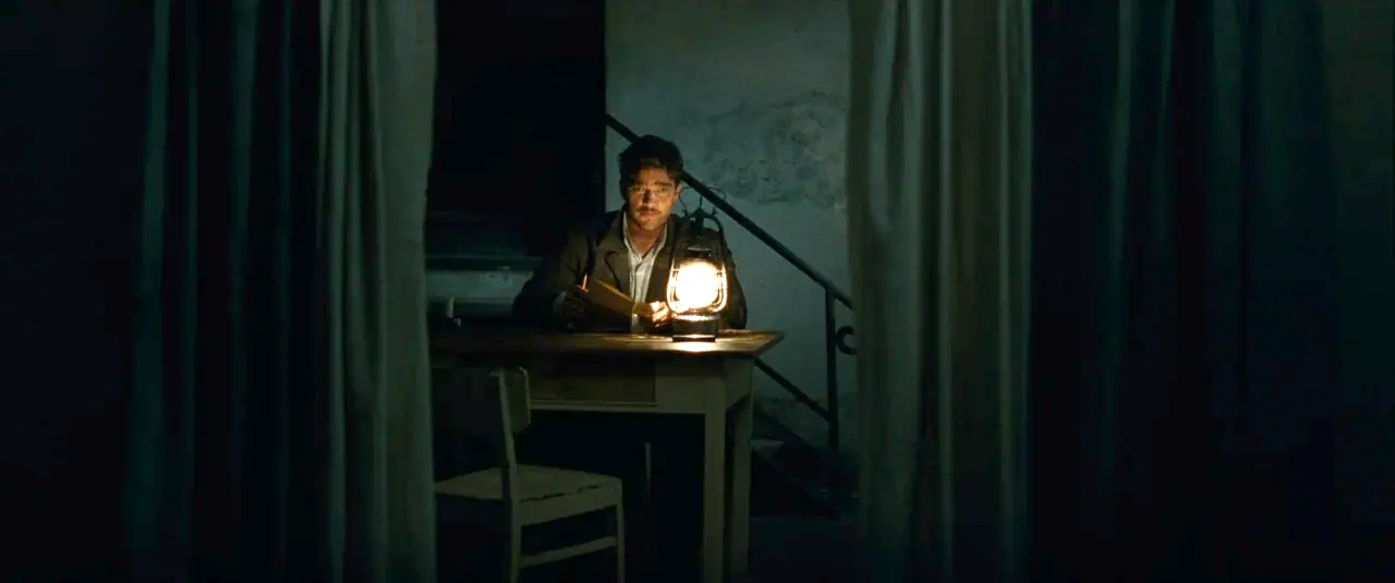 Phoenix (2014) movie still: medium — A man sits at a plain wooden table in a dimly lit, shadowed room. Curtains frame the foreground, while a stone…