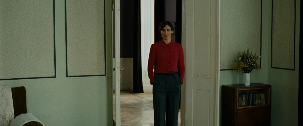 Phoenix (2014) movie still: fullbody — A room with pale green walls featuring framed panels stands adjacent to a hallway. A woman in a red shirt…