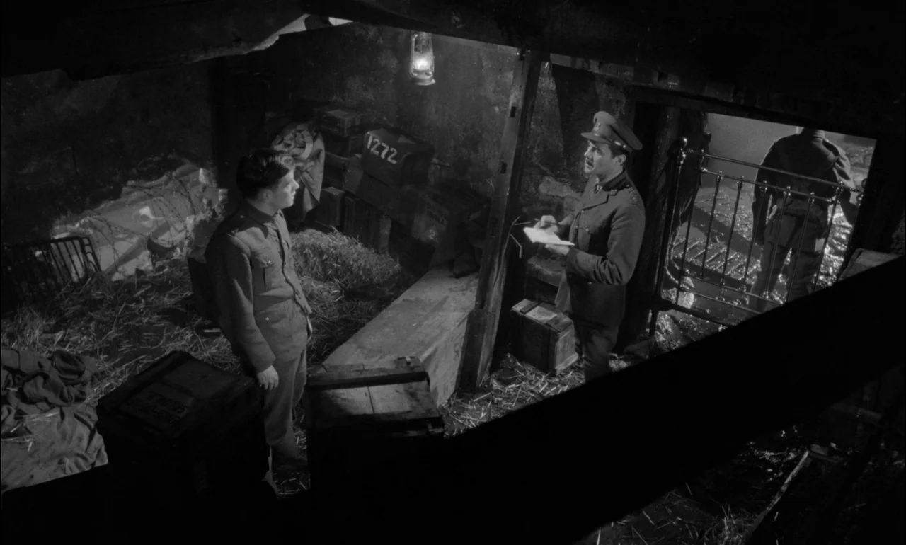 King & Country (1964) movie still: wide — A rustic bunker interior is filled with straw, wooden crates, and military supplies. An officer in uniform…