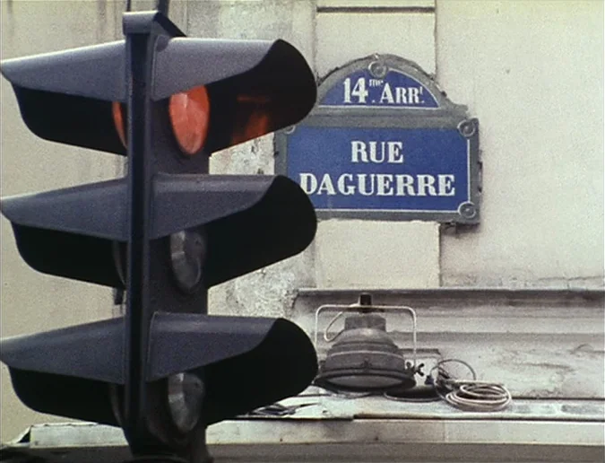 Daguerréotypes (1976) movie still: closeup — A Parisian street corner sign for Rue Daguerre sits above an industrial lamp. A large traffic light in…