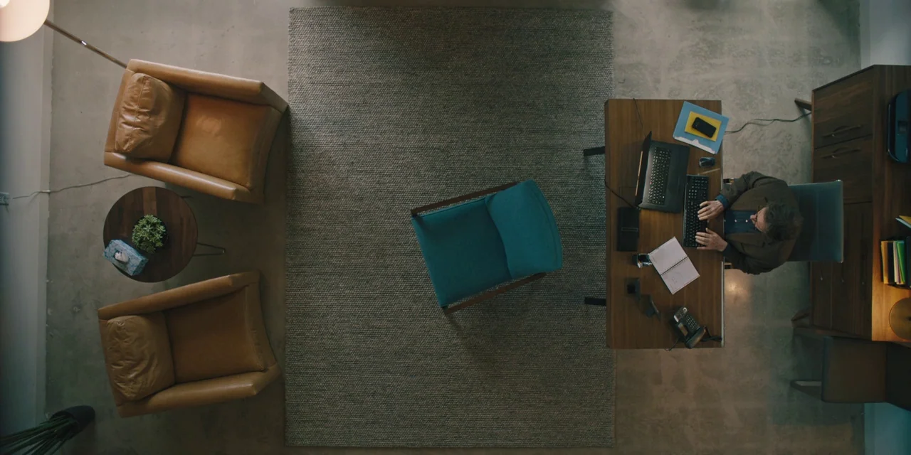 Hereditary (2018) movie still: wide — A top-down view of a modern professional office with leather chairs and wooden furniture. A man works at a…