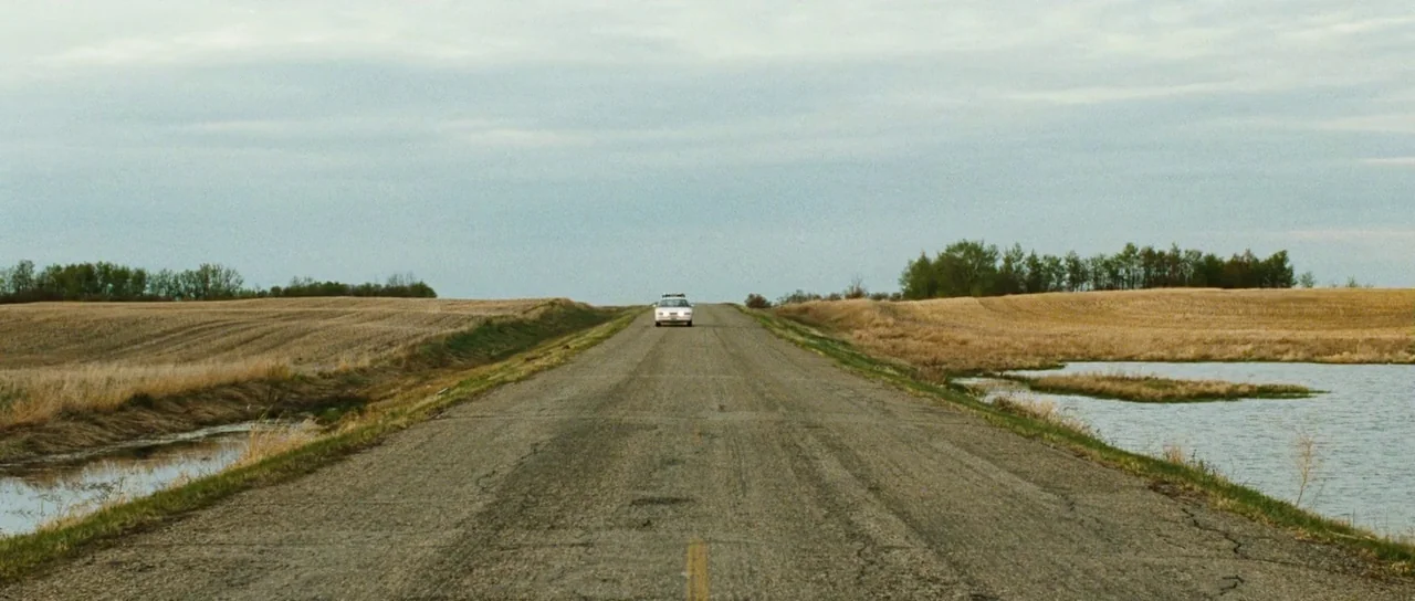 Surveillance (2008) movie still: wide — A long, straight paved road cuts through flat, harvested farmland under a cloudy sky. A white sedan travels…