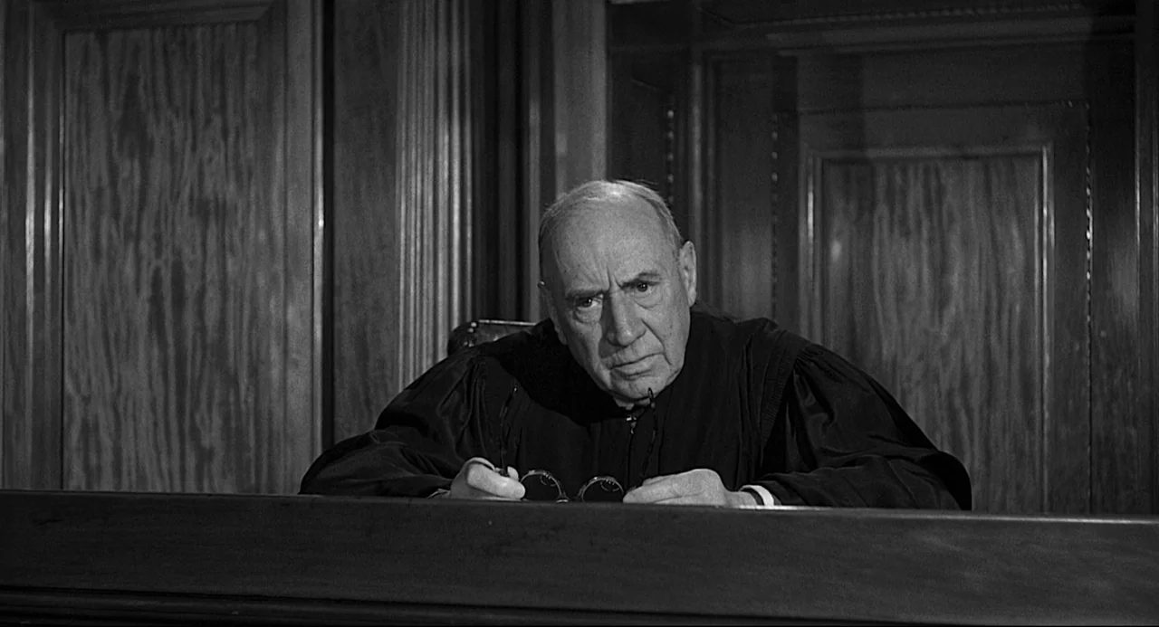 Anatomy of a Murder (1959) movie still: medium — A courtroom features dark wooden paneling behind a raised judicial bench. An elderly judge in a…