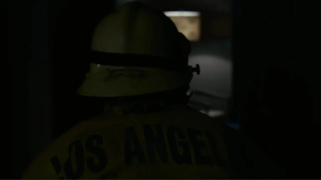 Quarantine (2008) movie still: medium — A dark, shadowed indoor space viewed over the shoulder of a person in uniform. The person wearing a fire…