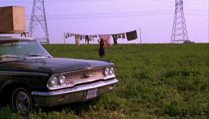 Highway 61 (1991) movie still: wide — An open green field contains a clothesline and large metal utility towers. A woman walks toward the laundry…
