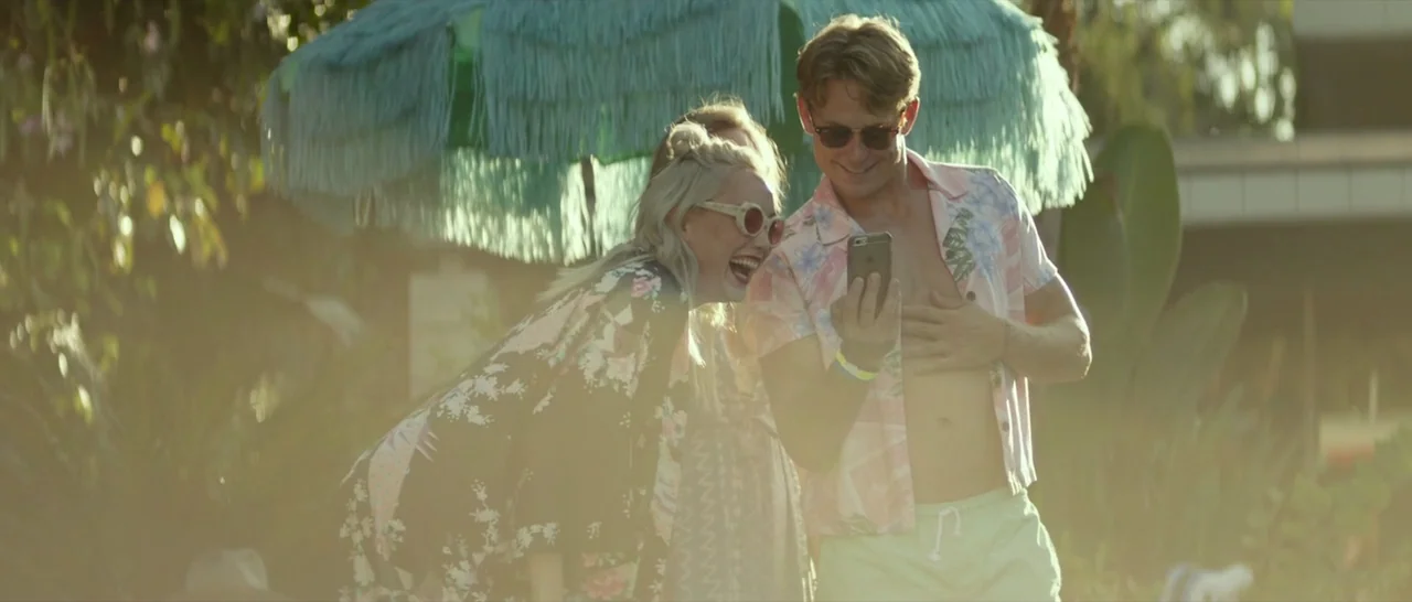 Ingrid Goes West (2017) movie still: medium — A sunny poolside setting with a blue tasseled umbrella visible in the blurred background. A young man…
