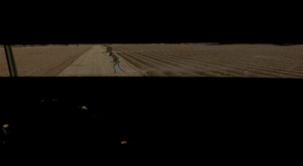 Near Dark (1987) movie still: wide — A vast, flat rural field is divided by a dirt track under a wide sky. A lone figure in light clothing moves…
