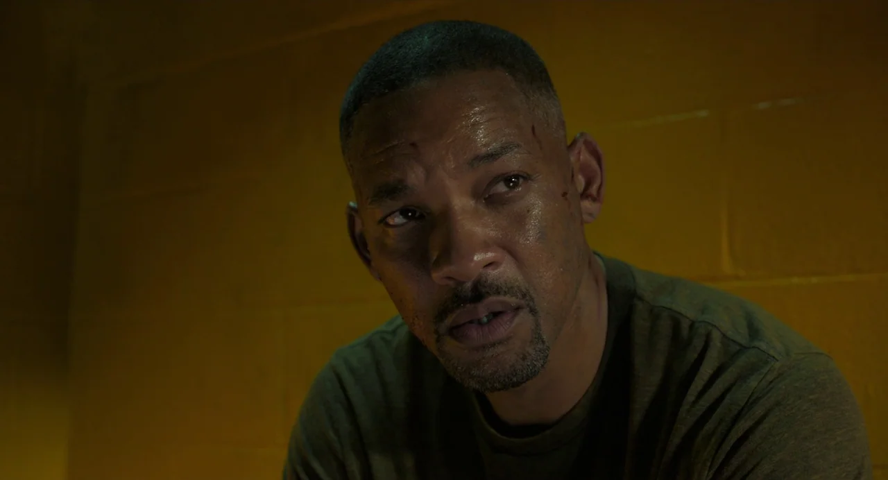 Gemini Man (2019) movie still: closeup — A man stands against a mustard-colored painted cinder block wall in a dimly lit room. He gazes towards the…