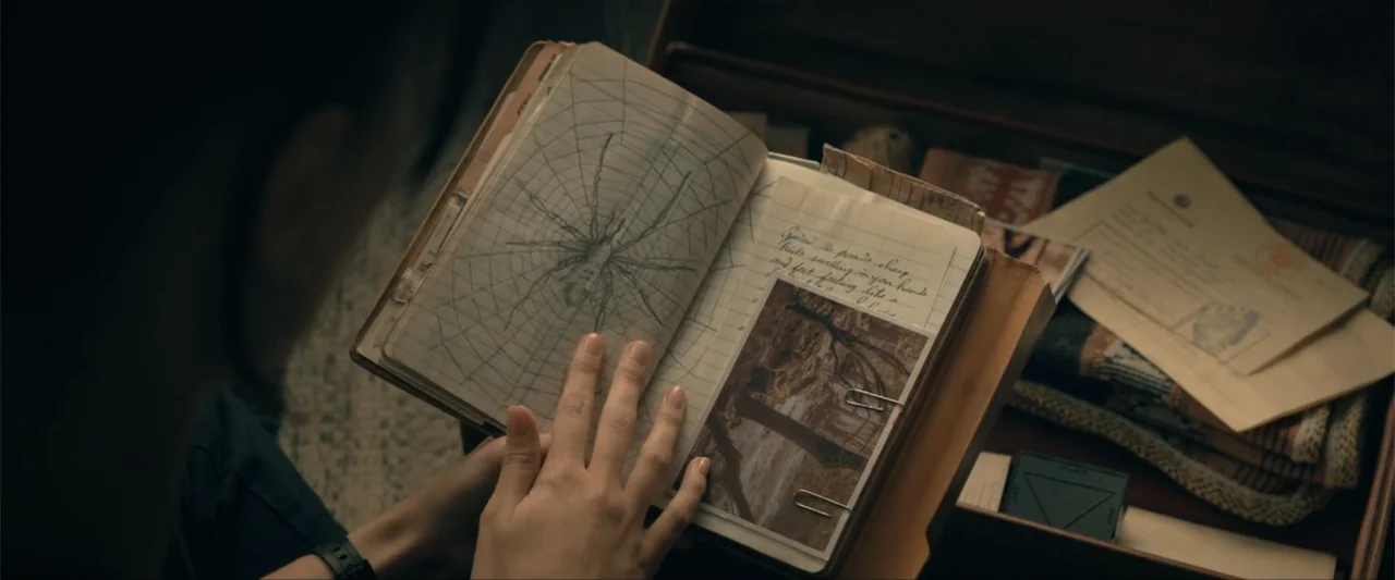 Madame Web (2024) movie still: closeup — A notebook lies open in a person's hands inside a cluttered wooden storage trunk. The left page features a…