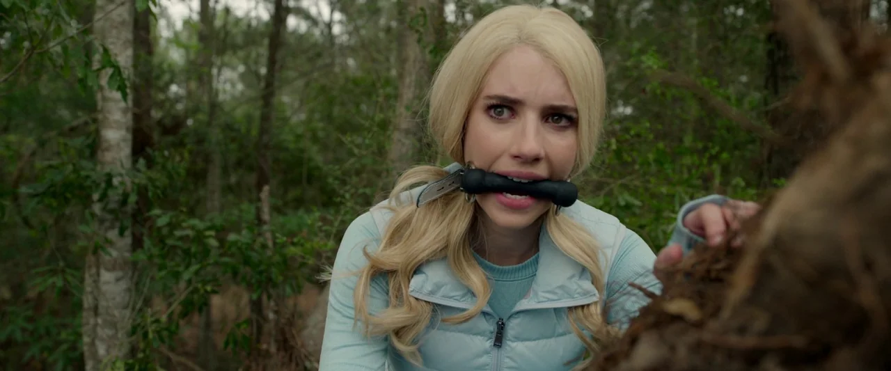 The Hunt (2020) movie still: medium — A dense patch of woodland with blurry green foliage and tree trunks in the background. A young woman with…