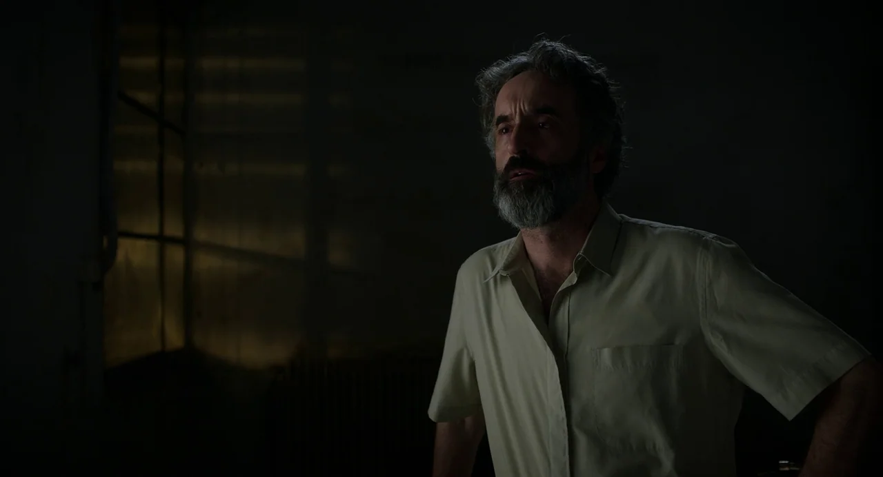 Crimes of the Future (2022) movie still: medium — A dark, sparsely furnished interior is defined by low light from a nearby window. A man with a…