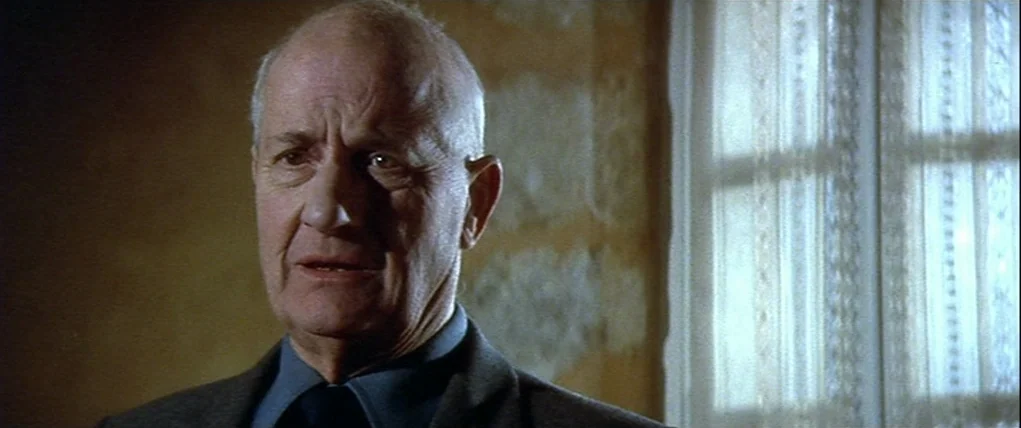 Love Unto Death (1984) movie still: closeup — A man stands indoors near a window covered by a white lace curtain. The wall behind him has a yellow,…