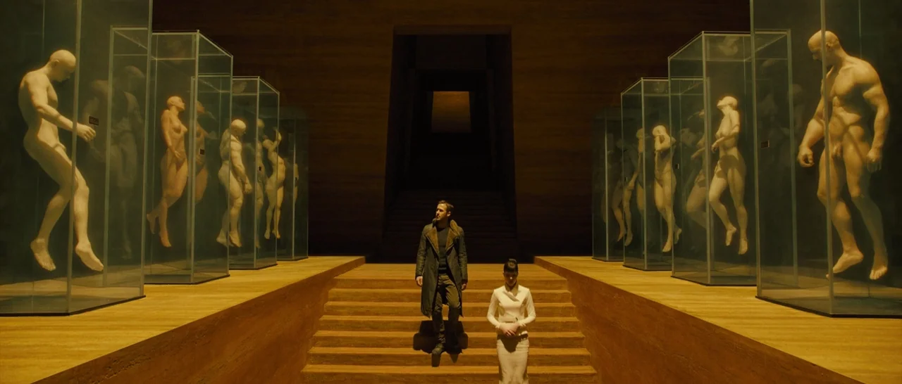 Blade Runner 2049 (2017) movie still: wide — A dark, expansive minimalist chamber features parallel rows of large glass pillars enclosing human…