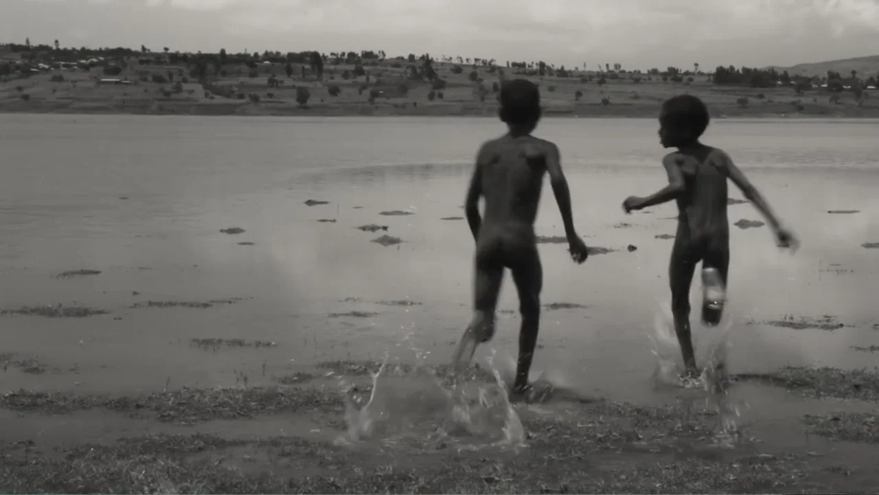 Faya Dayi (2021) movie still: fullbody — A rural lakeshore features a body of water and a grassy bank under clouds. Two children run into the water,…