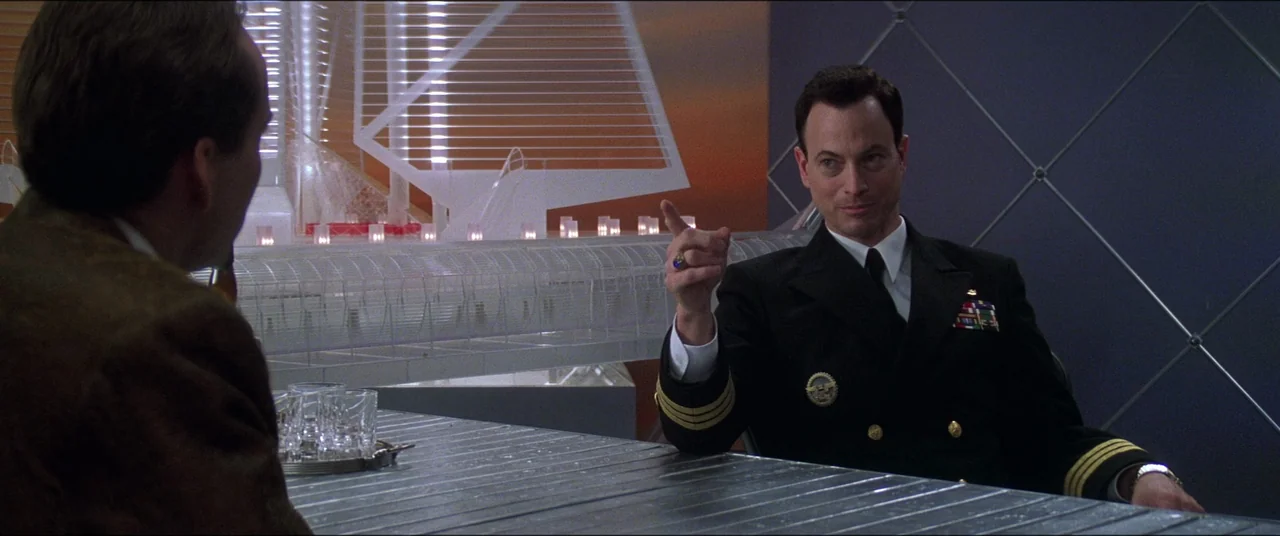 Snake Eyes (1998) movie still: medium — A modern, minimalist office featuring a large metallic table and an architectural model background. A…