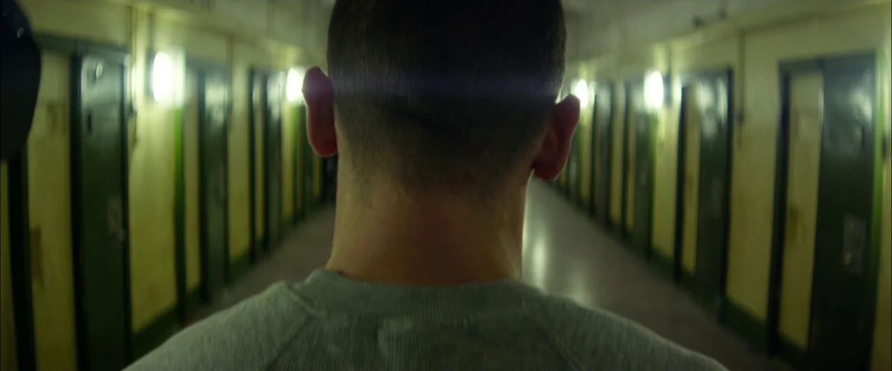 Starred Up (2013) movie still: medium — A long, symmetrical prison hallway lined with numerous green metal doors on both sides. A man is seen from…