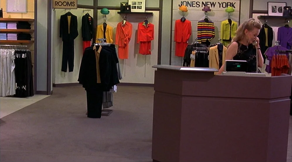 Jackie Brown movie still: wide — A quiet retail clothing shop features racks of colorful suits and wall-mounted garment displays. A woman stands…