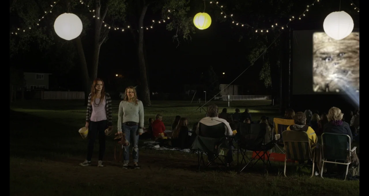 A.R.C.H.I.E. (2016) movie still: wide — A grassy park at night features a movie projector screen and rows of seated viewers. Two young women stand…