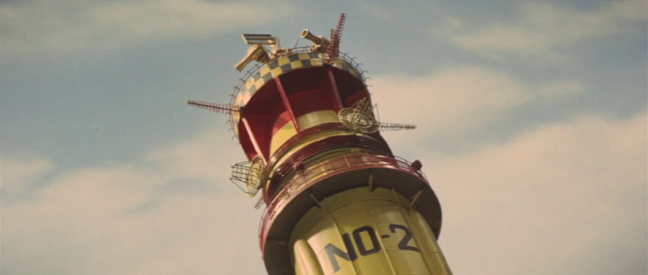 Son of Godzilla (1967) movie still: wide — A tall cylindrical tower labeled NO-2 rises against a cloudy sky. The structure features red and yellow…