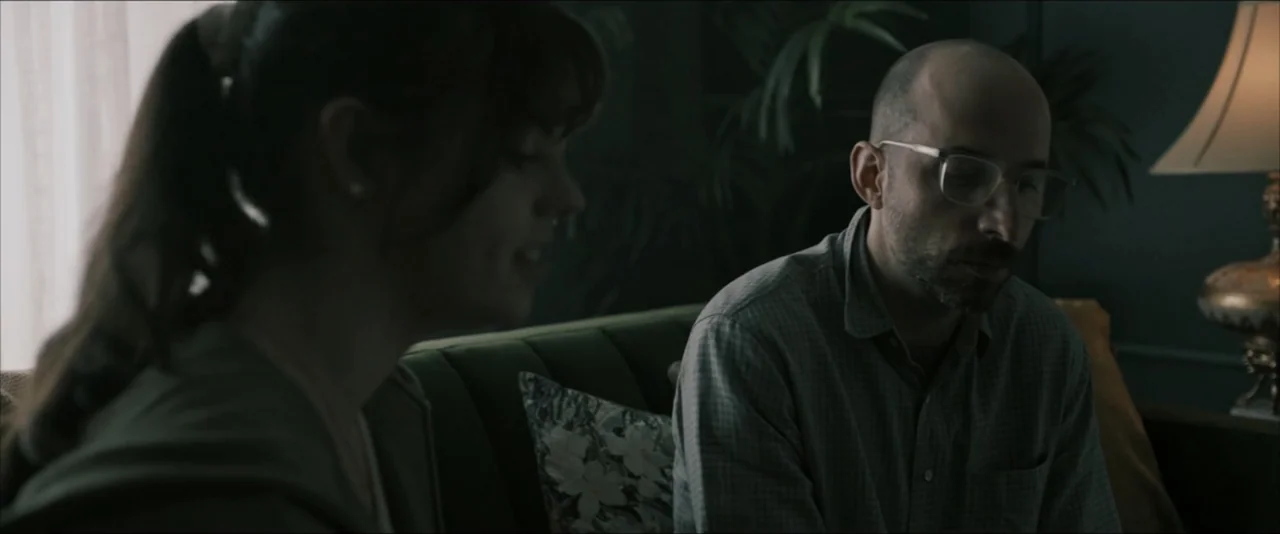 It Feeds (2025) movie still: medium — An interior living room with deep green walls and an ornate lamp on the side. A woman in a dark hoodie sits…