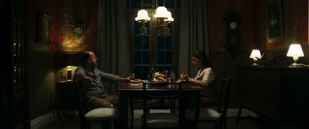 When Evil Lurks (2023) movie still: wide — A dimly lit room features patterned red wallpaper and wooden furniture with vintage accents. A man and a…