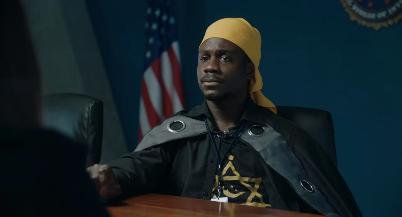 The Day Shall Come (2019) movie still: medium — An interior office space features a dark wall adorned with an American flag. A man wearing a yellow…