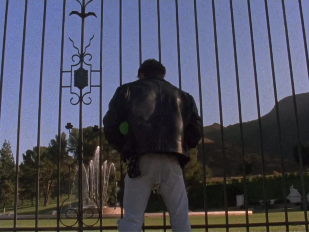 The Living End (1992) movie still: medium — A public park area seen through an ornate black metal gate at dusk. A man in a leather jacket stands at…