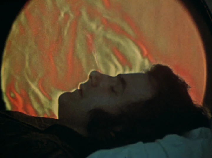 Lucifer Rising (1972) movie still: closeup — A man lies with his eyes closed against a large, glowing orange circular screen. The warm light from…