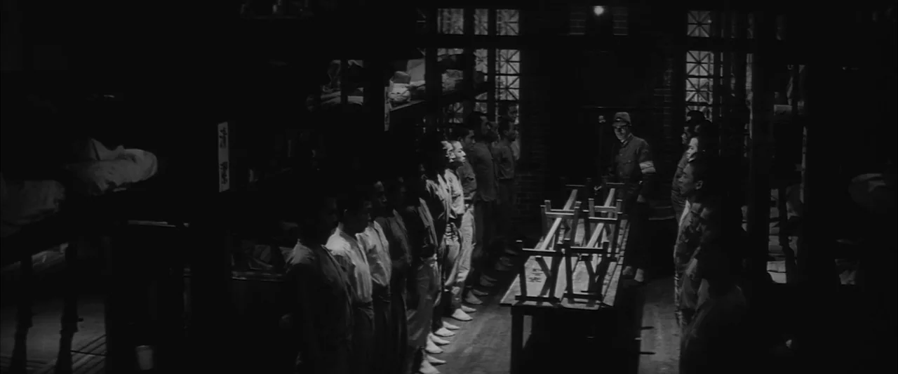 The Human Condition II: Road to Eternity (1959) movie still: wide — A dimly lit room with bunk beds and rows of wooden stools aligned.