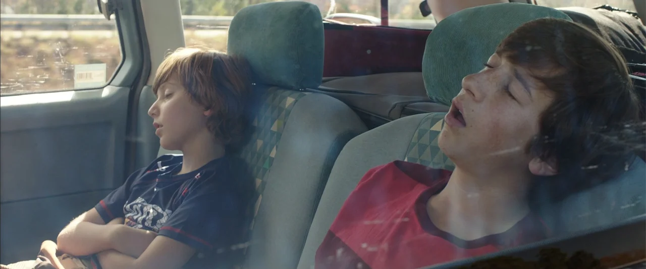 Vacation (2015) movie still: medium — The interior of a moving car shows two children sleeping in patterned fabric seats. The sunlight reflects off…