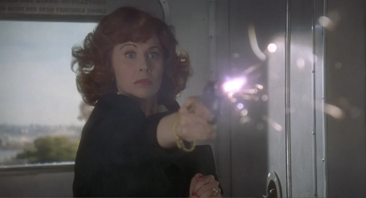 Fatal Instinct (1993) movie still: medium — The interior of a train car features a large side window and industrial paneling. A woman aims a small…