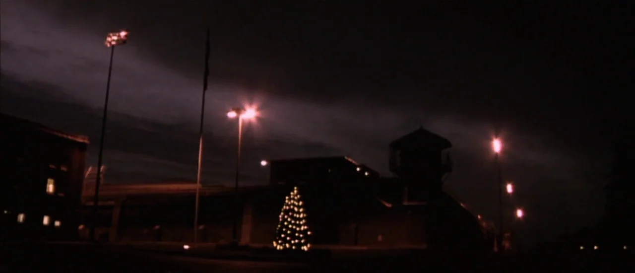 Dancer in the Dark (2000) movie still: establishing — A prison facility at night features a watchtower and a decorated Christmas tree. The grounds…
