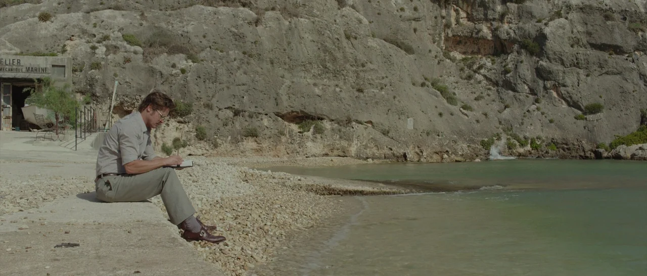 By The Sea (2015) movie still: wide — A man sits on a concrete ledge next to a calm inlet near cliffs. He concentrates on writing in a small book…