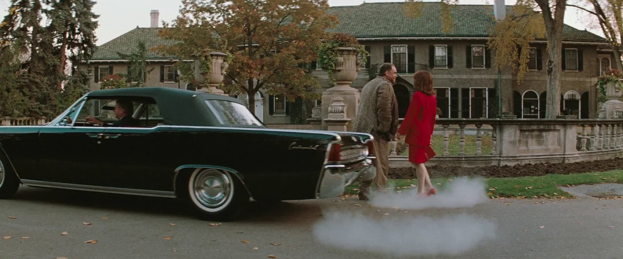The Adjuster (1991) movie still: wide — A stone mansion with a low wall sits behind a paved driveway with autumn trees. A man and a woman stand near…