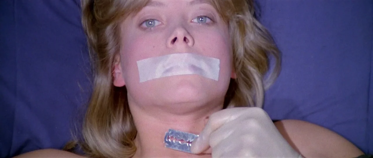 The New York Ripper (1982) movie still: closeup — A close-up of a blonde woman lying on a dark blue surface. A gloved hand holds a razor blade…