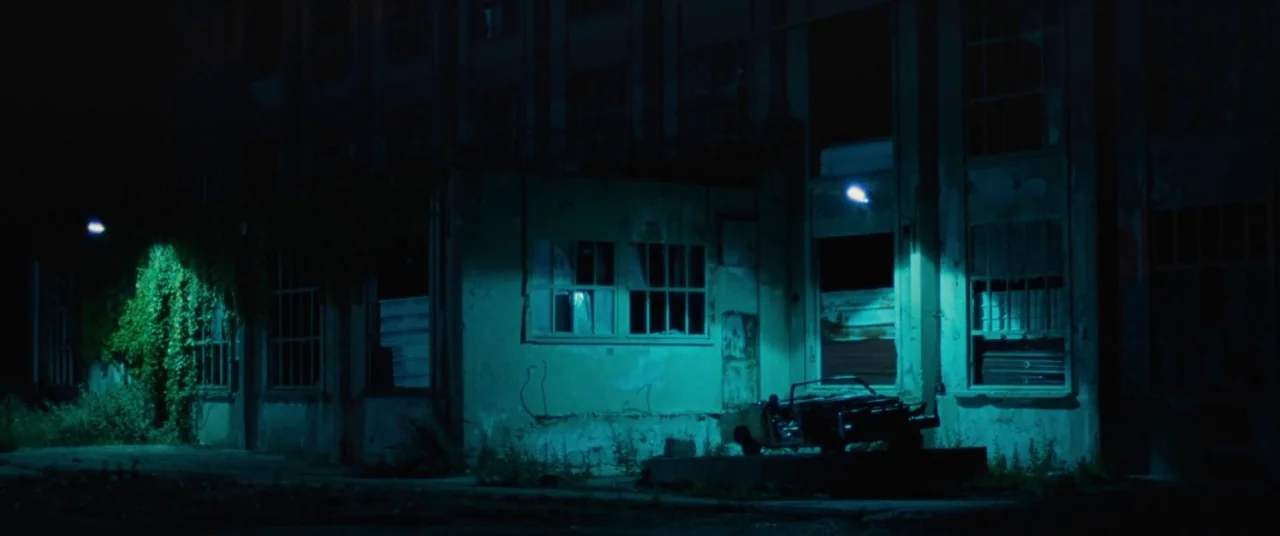 Knife+Heart (2018) movie still: wide — An abandoned industrial building with shattered windows sits in total darkness at night. A derelict car is…